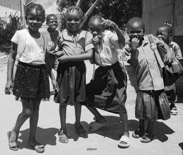 Eine Gruppe lachender Kinder, die nebeneinander auf einem Bürgersteig stehen, mit Gebäuden, Bäumen und Pflanzen im Hintergrund und einem klaren blauen Himmel, dargestellt in Schwarz-Weiß.