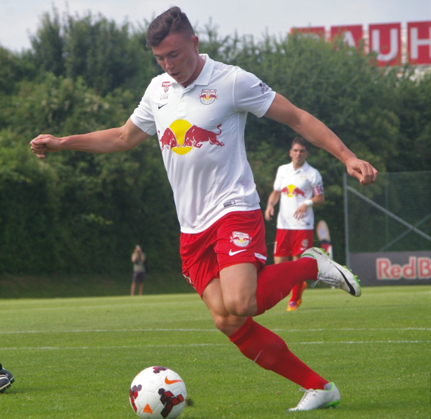 Ein professioneller Fussballspieler, der einen Ball auf einem grünen Feld kickt, mit "RB Leipzig" im Hintergrund und einem klaren blauen Himmel.