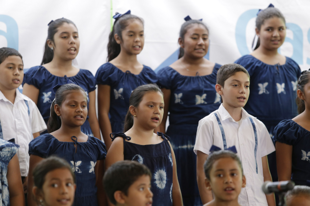 Gruppe von Kindern, die vor einem Mikrofon singen, mit einem Schulchor-Banner im Hintergrund.