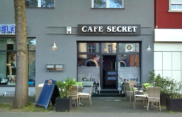 Ein Café in Berlin, Deutschland, mit einem Gebäude mit Fenstern und einer Tür, einem Schild mit Text, Außenbestuhlung mit Stühlen und Tischen, Topfpflanzen, einer Tafel, einem Baum und Gras.