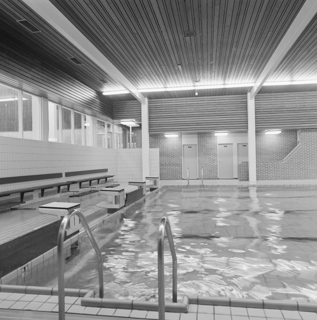 Große Hallenbad mit gefliestem Boden, Metallstangen, Bänken, Wänden, Türen, Lampen und Fenstern, gef\u00fllt mit klarem Wasser, Decke oben sichtbar.