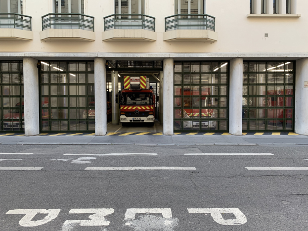 Feuerwache mit vor dem Gebäude geparktem Feuerwehrauto, mit Fenstern, Geländern, Säulen, Glastüren und einer Straße unten.