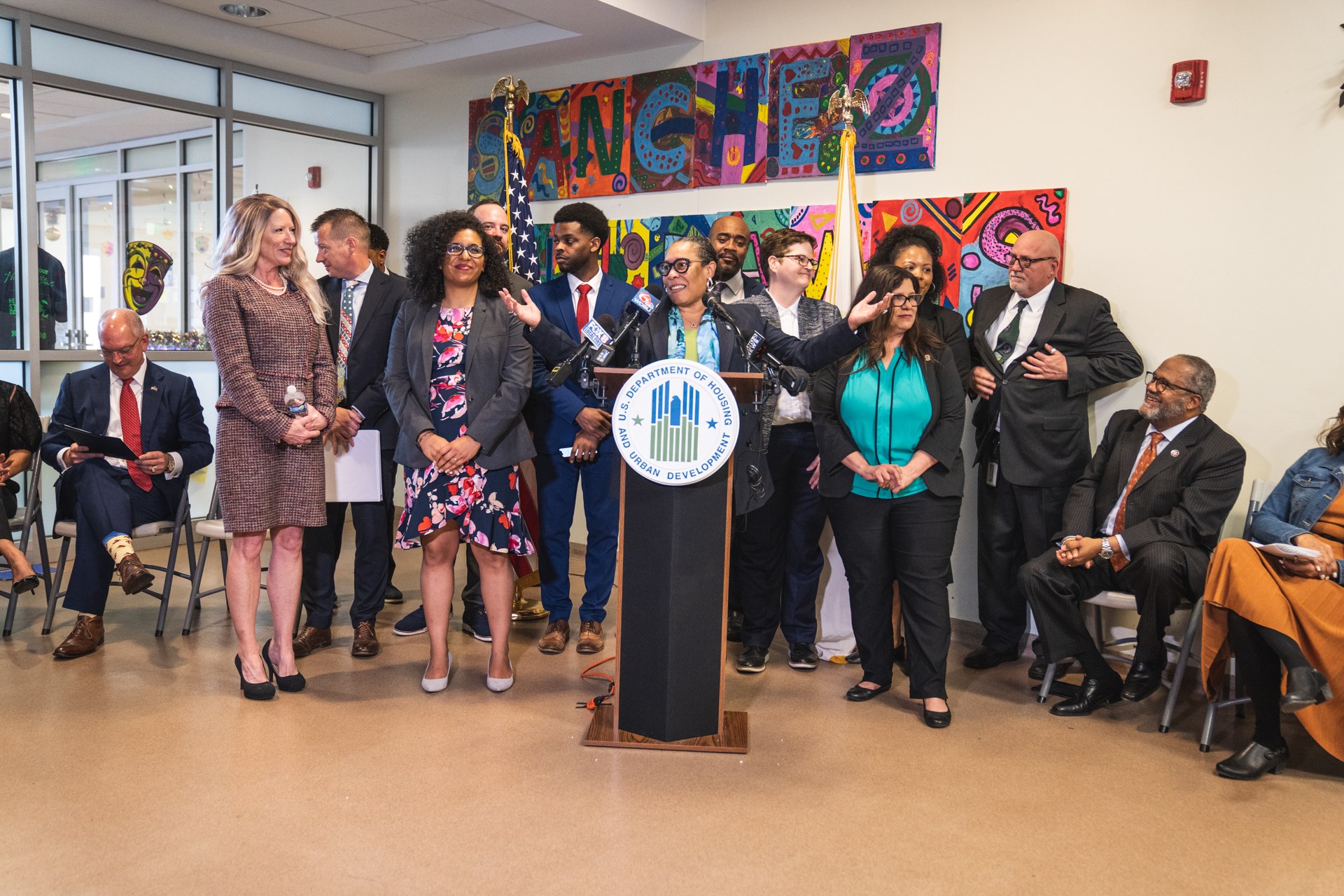 Gruppe von Menschen vor einem Podium mit Mikrofon und einer Tafel mit Text, einige sitzen auf Stühlen und andere stehen, mit Flaggen, einer Tafel, einem Feuerlöscher und Deckenlampen an der Rückwand.