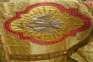 Nahaufnahme eines Priestergewandes mit goldenen und roten Stickereien in komplexen Mustern vor einer Wand im Hintergrund.