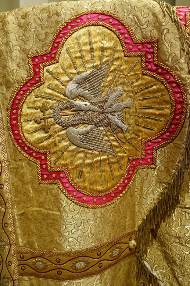 Nahaufnahme eines Priestergewandes mit goldenen und roten Stickereien in komplexen Mustern vor einer Wand im Hintergrund.