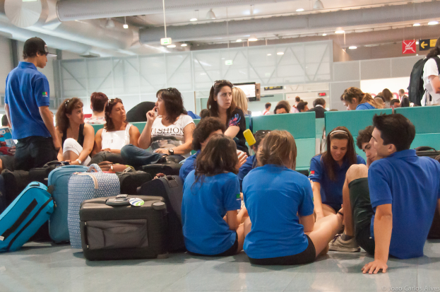 Ein belebter Flughafeninnenraum mit vielen Menschen, die auf Flüge warten, Gepäck links und Glastüren mit Richtungsschildern im Hintergrund.