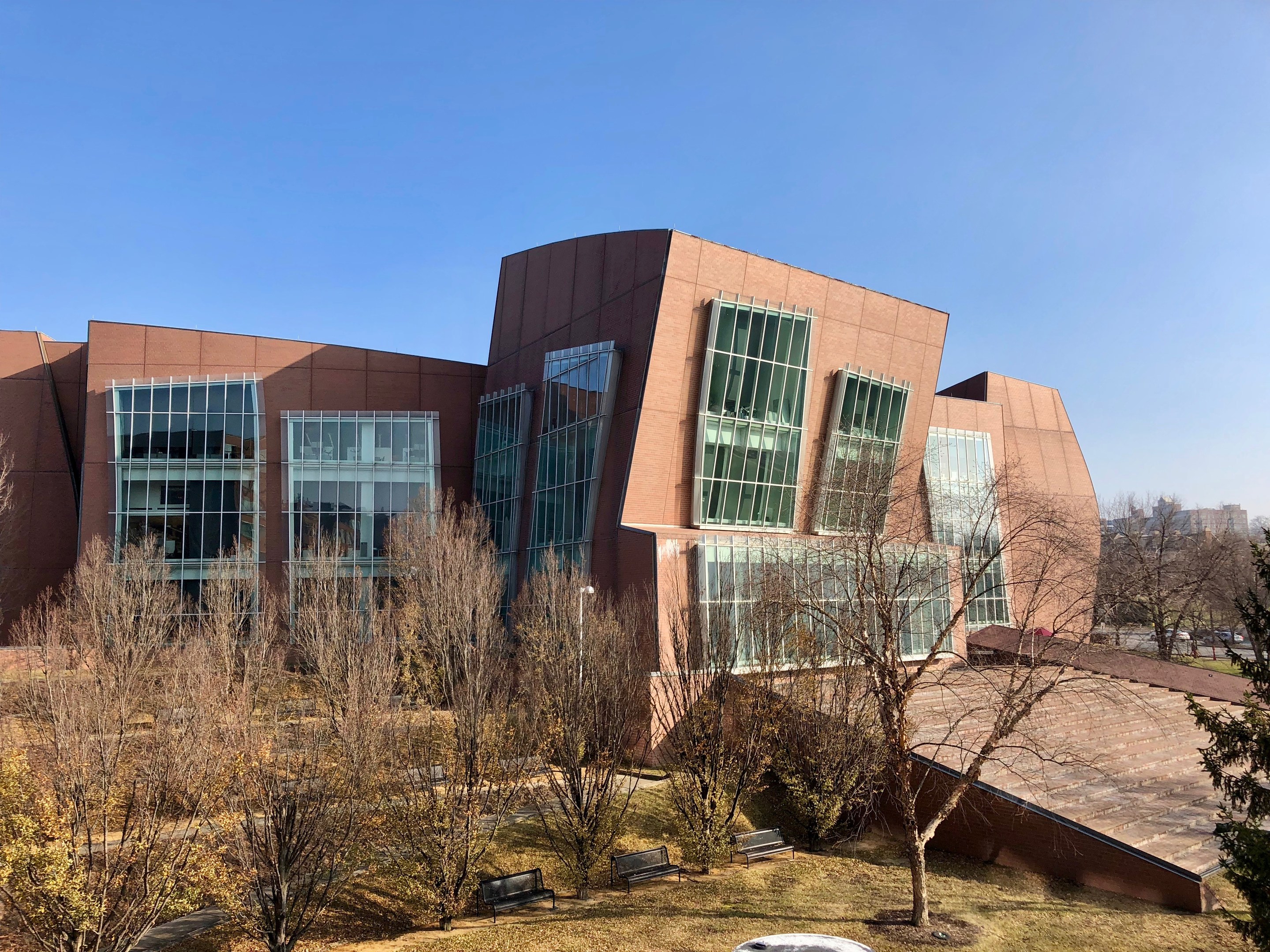 Großes mehrstöckiges Gebäude mit zahlreichen Fenstern, umgeben von Bäumen, Bänken, Gras und einem Laternenmast, mit anderen Gebäuden und einem klaren blauen Himmel im Hintergrund, identifiziert als der neue Campus der University of Illinois at Chicago.