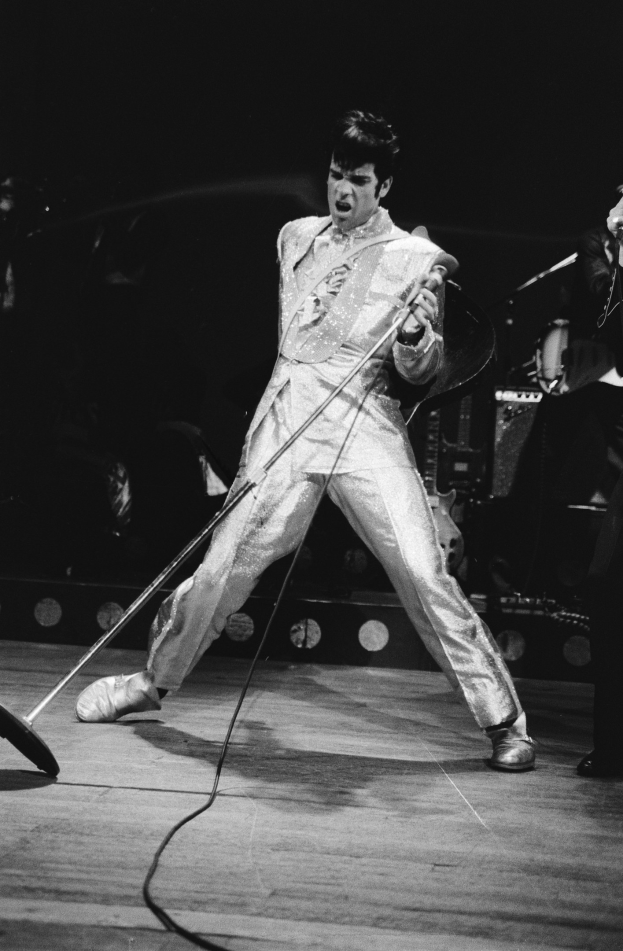 Elvis Presley in einem weißen Jumpsuit performs auf der Bühne des Apollo Theaters in Las Vegas, hält ein Mikrofon in der Hand und hat einen anderen Musiker rechts von sich und Bühneninstrumente und Lichter im Hintergrund.