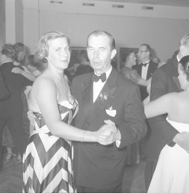 Ein Mann in einem schwarzen Blazer, weißem Hemd und schwarzen Hosen tanzt mit einer Frau in einem unbekannten Outfit bei einem formellen Ereignis, indem sie sich an den Händen halten und zur Musik schaukeln.