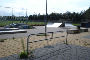 Ein Skatepark mit Rampen, Geländern, Grünflächen, Pfosten, Bäumen, Gebäuden und einem bewölkten Himmel im Hintergrund.