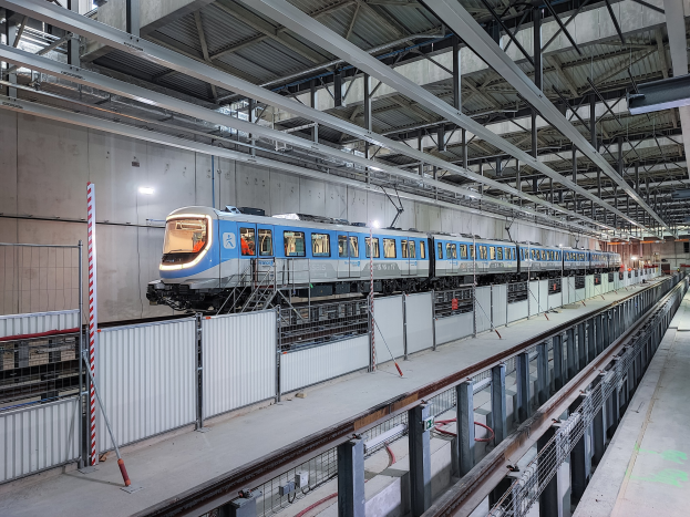 Ein blauer und weißer Zug fährt durch eine große Indoor-Station mit einer Plattform auf der rechten Seite und einer Wand im Hintergrund, unter einer Decke mit Stangen und Lampen.