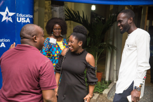 Gruppe von Menschen, die vor einem blauen Zelt mit einem Banner, Topfpflanzen, einer Säule und einem Dach mit Deckenleuchten sprechen.