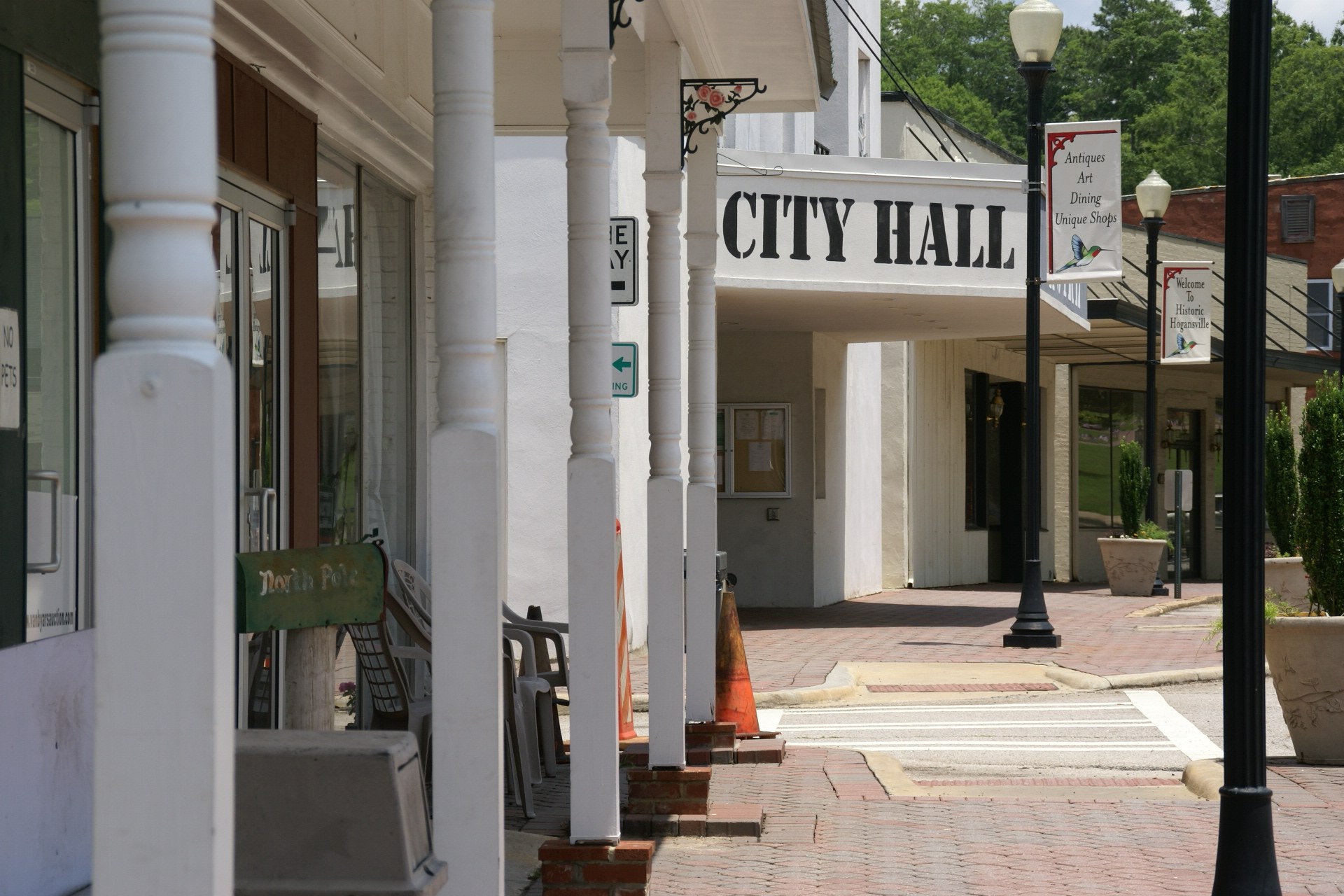 Ein Rathaus umgeben von Gebäuden, Straßenlaternen, Straßenschildern, Fahrzeugen, Verkehrskegeln, Stühlen, Topfpflanzen, Bäumen und einem bewölkten Himmel.