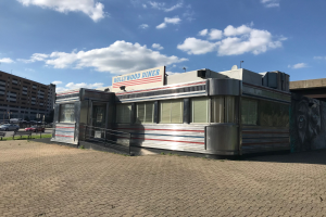 Außenansicht des Hollywood Diners mit seiner Leuchtschrift, das sich auf einer Straße befindet, die von Gebäuden, Straßenlaternen und Fahrzeugen gesäumt ist, unter einem bewölkten Himmel.