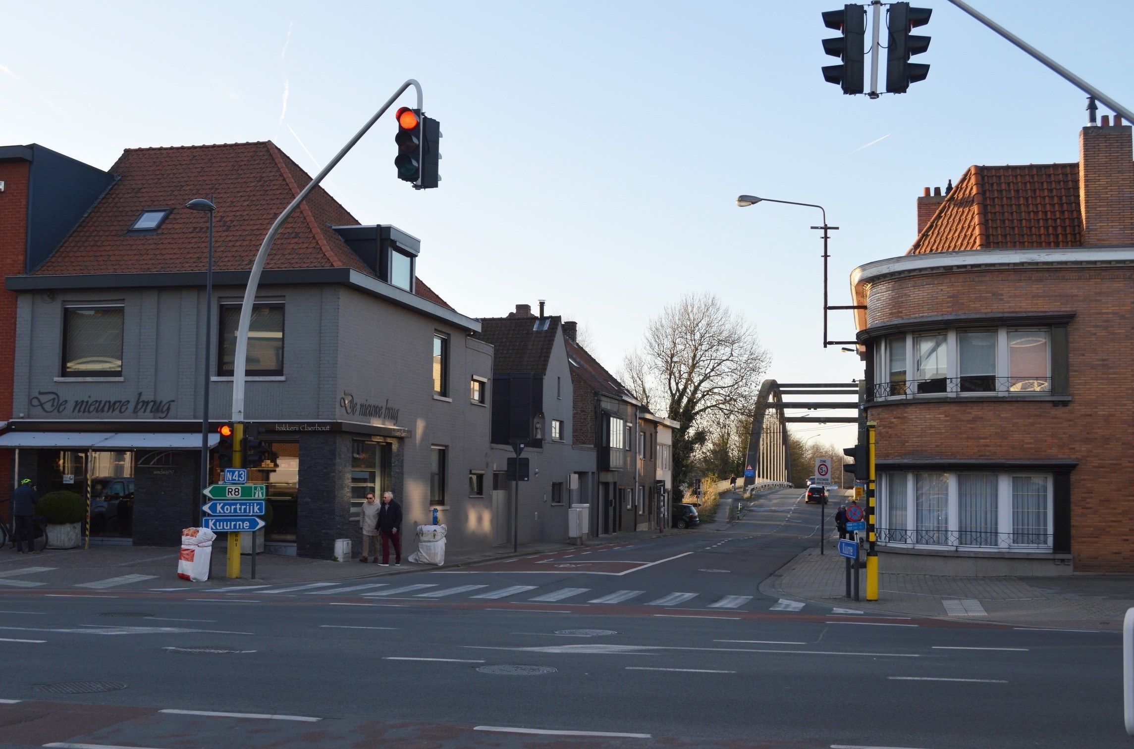 Stadtstraßenkreuzung mit Ampel, Fußgängern, Fahrrädern, Bäumen, Gebäuden, Straßenschildern, Laternen, einem Bogen und einem klaren blauen Himmel.