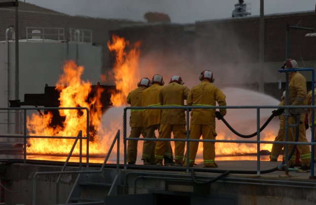 Eine Gruppe von Feuerwehrleuten in Helmen steht auf dem Dach eines Gebäudes und hält Rohre, mit Geländern, Treppen, einem Gebäude und dem Himmel im Hintergrund.