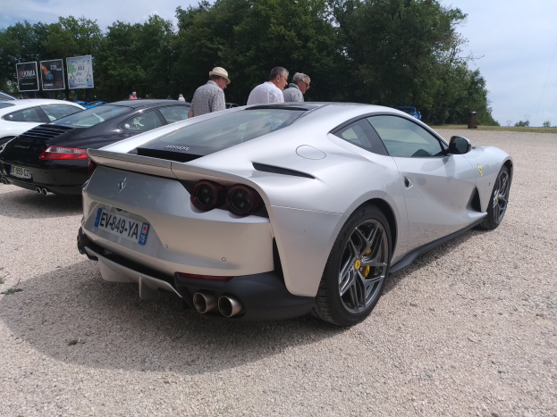 Ein roter Ferrari 812 Superfast bei der Goodwood Festival of Speed, umgeben von Menschen mit Mützen, Bäumen, Texttafeln und einem bewölkten Himmel.