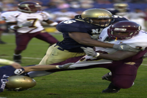 Zwei Fußballspieler in Helmen und Uniformen ringen auf dem Feld, während andere im Hintergrund zuschauen.