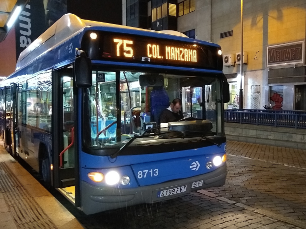 Ein blauer Bus parkt nachts auf einer Straße mit einer Person darin, einer Handtasche im Vordergrund und Gebäuden, Schildern, Geländern und Lichtern im Hintergrund.