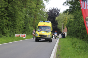Ambulanz fährt auf einer Straße mit Radfahrern äquivalent, Gras und Bäume auf beiden Seiten, Häuser, Mäste und einen klaren blauen Himmel im Hintergrund.