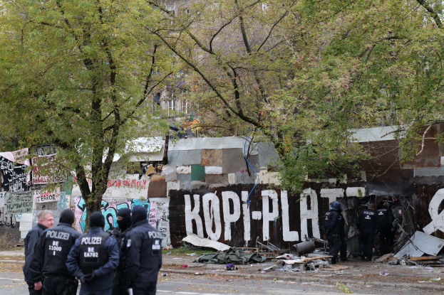 Eine Gruppe von Polizisten in schwarzen Uniformen und Helmen steht vor einem mit Graffiti bedeckten, baufälligen Gebäude mit verstreuten Trümmern und Bäumen im Hintergrund.