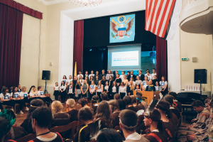 Eine Gruppe von Menschen steht an einem Podium mit einem Mikrofon, flankiert von Fahnen und Lautsprechern, und spricht vor einem sitzenden Publikum während eines kulturellen Ereignisses, mit einem Bildschirm, Vorhängen und einem Kronleuchter im Hintergrund.