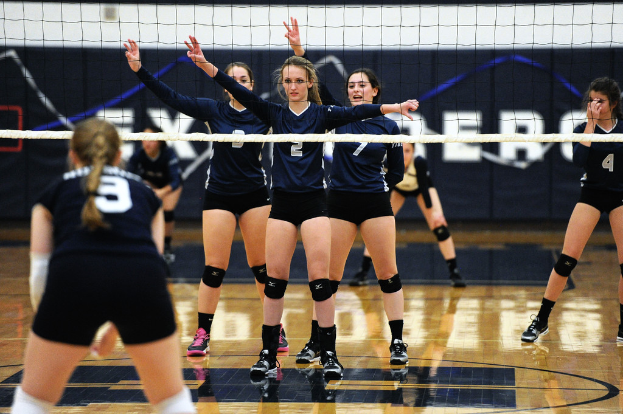 Drei Mädchen bei einem Volleyballspiel auf dem Feld stehend, ein Netz vor ihnen und ein Zaun im Hintergrund.