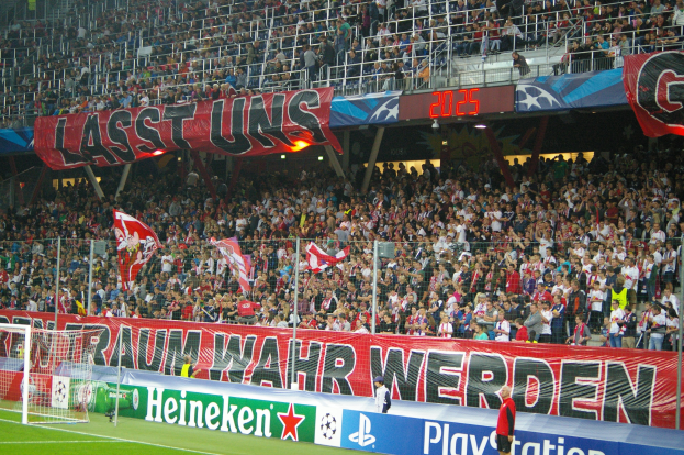 Fußballspiel in einem Stadion mit großer Zuschauermenge, saftig grünem Rasen, Torpfosten links, Bodenbanner, Menschen mit Fahnen und Hintergrundgeländer, Tafeln, Lichter und andere Gegenstände.