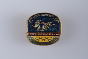 Ein Abzeichen mit dem Logo der Olympischen Spiele 1980, das zwei Läufer und den Text "Olympische Spiele" darüber zeigt.