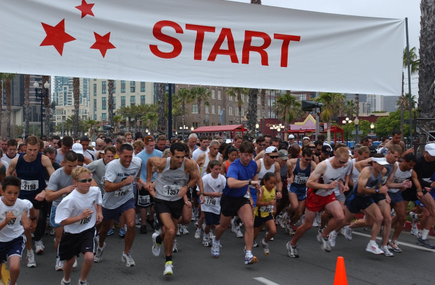 Gruppe von Menschen beim Marathon mit einem Verkehrskegel im Vordergrund und einem Banner im Hintergrund, umgeben von Bäumen, Laternenmasten, Gebäuden und einem klaren blauen Himmel.