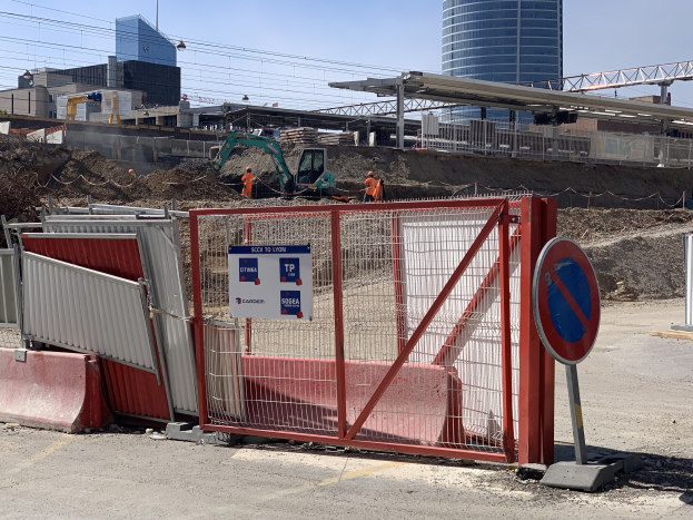 Baustelle mit einem roten Tor, Metallzaun, Gebäuden, Kränen, Masten, Drähten und Arbeitern, unter einem klaren blauen Himmel.