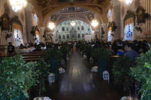 Eine Hochzeitszeremonie in einer Kirche mit sitzenden und stehenden Gästen, dekoriert mit Zimmerpflanzen, Skulpturen, Vorhängen, Kronleuchtern und einem Bildschirm im Hintergrund.