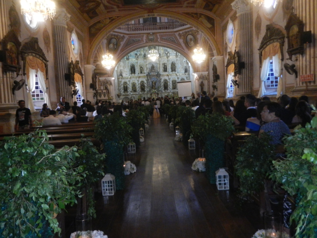 Eine Hochzeitszeremonie in einer Kirche mit sitzenden und stehenden Gästen, dekoriert mit Zimmerpflanzen, Skulpturen, Vorhängen, Kronleuchtern und einem Bildschirm im Hintergrund.