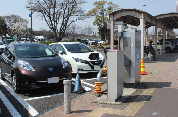 Ladestation für Elektroauto in Japan mit Autos auf der Straße, Verkehrskegeln, einer Person auf dem Gehweg und umgebender Infrastruktur gegen einen Himmel als Hintergrund.
