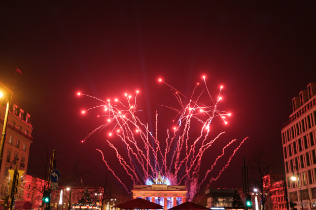 Eine städtische Straße voller Gebäude, Bäume, Laternen, Verkehrszeichen, Zelte und Menschen, mit einem Feuerwerk am Himmel während des Silvesterfeierns in Berlin.