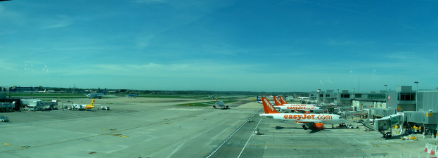 Eine Flughafen-Szene mit Flugzeugen, Gebäuden, Polen, Fahrzeugen und Bäumen, mit einem Himmel darüber und einer Straße darunter und einem Zaun im Vordergrund.