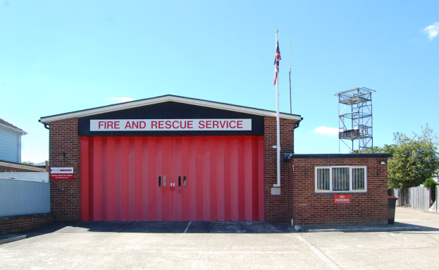 Feuerwehr- und Rettungsdienstgebäude mit roter Tür, Fenstern, einem Namensschild, einer Flaggenstange mit einer Flagge, einem Metallturm, einem Zaun, Bäumen und einem bewölkten Himmel.