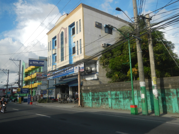 Stadtstraßenszene mit Gebäuden, Fahrzeugen, Straßenlaternen, Oberleitungen, Namensschildern, Bäumen und bewölktem Himmel, mit einem im Vordergrund stark mit Kabeln bedeckten Gebäude.