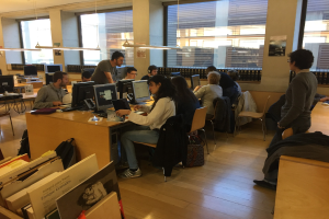 Gruppe von Menschen, die an Computern auf Schreibtischen in einer Bibliothek mit Bücherregalen, Monitoren, Tastaturen, Mäusen, Taschen auf dem Boden, Deckenlampen und Glasfenstern im Hintergrund arbeiten.