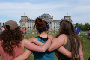 Drei Frauen in verschiedenen farbigen Kleidern und eine mit einer Mütze stehen vor dem Reichstagsgebäude in Berlin, Deutschland, mit Menschen, Fahnen, Bäumen, Wolken und dem Himmel im Hintergrund.