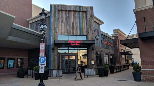 Außenansicht eines Restaurants mit einer Frontwerbung, umgeben von Gebäuden, Straßenmöbeln und Grünpflanzen unter einem klaren blauen Himmel.