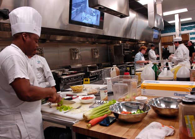 Eine Gruppe von Köchen in einer Küche beim Zubereiten von Speisen auf einem Schneidbrett, wobei ein Koch im Vordergrund einen weißen Kittel und eine Haube trägt und ein Messer in der Hand hält und von verschiedenen Utensilien, Schüsseln, Gemüse, einem Mobiltelefon, Flaschen und anderen Gegenständen umgeben ist, mit einem Fernseher und Deckenleuchten im Hintergrund.