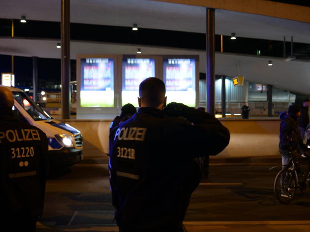 Eine Gruppe von Polizisten steht vor einem hell erleuchteten Gebäude in der Nacht, mit einem geparkten Auto links und einem Radfahrer rechts.