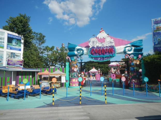 Karussell am Parkeingang mit Straße, eingezäuntem Bereich, Buden, Fahnen, Bäumen und bewölktem Himmel im Hintergrund.
