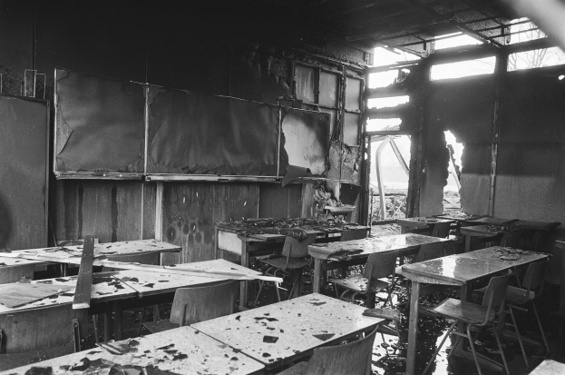 Schwarzes und weißes Foto eines verlassenen Klassenzimmers mit verbrannten Wänden, zerbrochenen Fenstern und verstreuten Tischen und Stühlen.