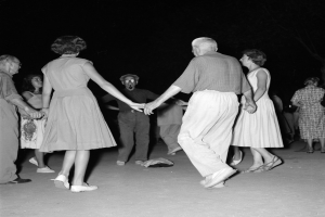 Eine Gruppe von Menschen tanzt nachts auf der Straße, hält sich an den Händen und ist von Mondlicht und Sternen beleuchtet, auf einem Schwarz-Weiß-Bild.