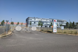 Ein großes Gebäude mit vielen Fenstern, identifiziert als First Solar, umgeben von einem Metallzaun, Bäumen, Pflanzen, Gras, Schildern, Straßenlaternen und einem Weg.