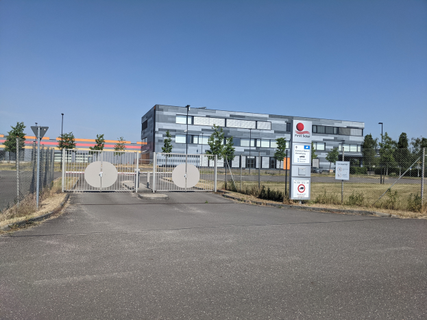 Ein großes Gebäude mit vielen Fenstern, identifiziert als First Solar, umgeben von einem Metallzaun, Bäumen, Pflanzen, Gras, Schildern, Straßenlaternen und einem Weg.