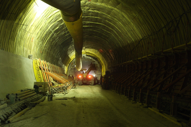 Baustelle mit einem großen zentralen Tunnel, Fahrzeugen, verstreuten hölzernen Objekten, Rohren und einer Wand auf der linken Seite, beleuchtet von Deckenlampen.
