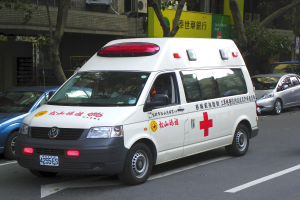 Ambulanz am Straßenrand mit anderen Fahrzeugen, Bäumen, Gebäuden, Schildern und Klimaanlagen im Hintergrund.